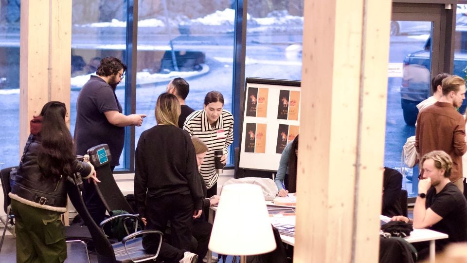 People gathered around a desk and whiteboard, leaning on the desk, working on something.